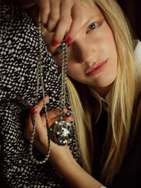 Close-up of a blond woman holding a beaded necklace with a spherical pendant, featuring a black and white patterned textile in the background.
