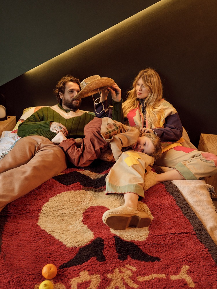 Family of three lies on a red and tan patterned rug, with a woman holding a cowboy hat over a child, and oranges nearby.