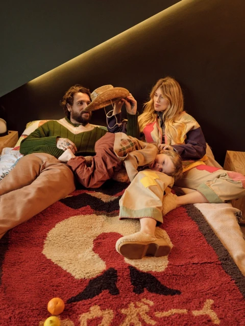 Family of three lies on a red and tan patterned rug, with a woman holding a cowboy hat over a child, and oranges nearby.