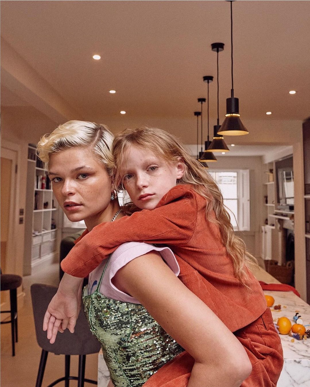 A young adult woman giving a piggyback ride to a young child in an indoor environment, with visible kitchen and interior decorations.