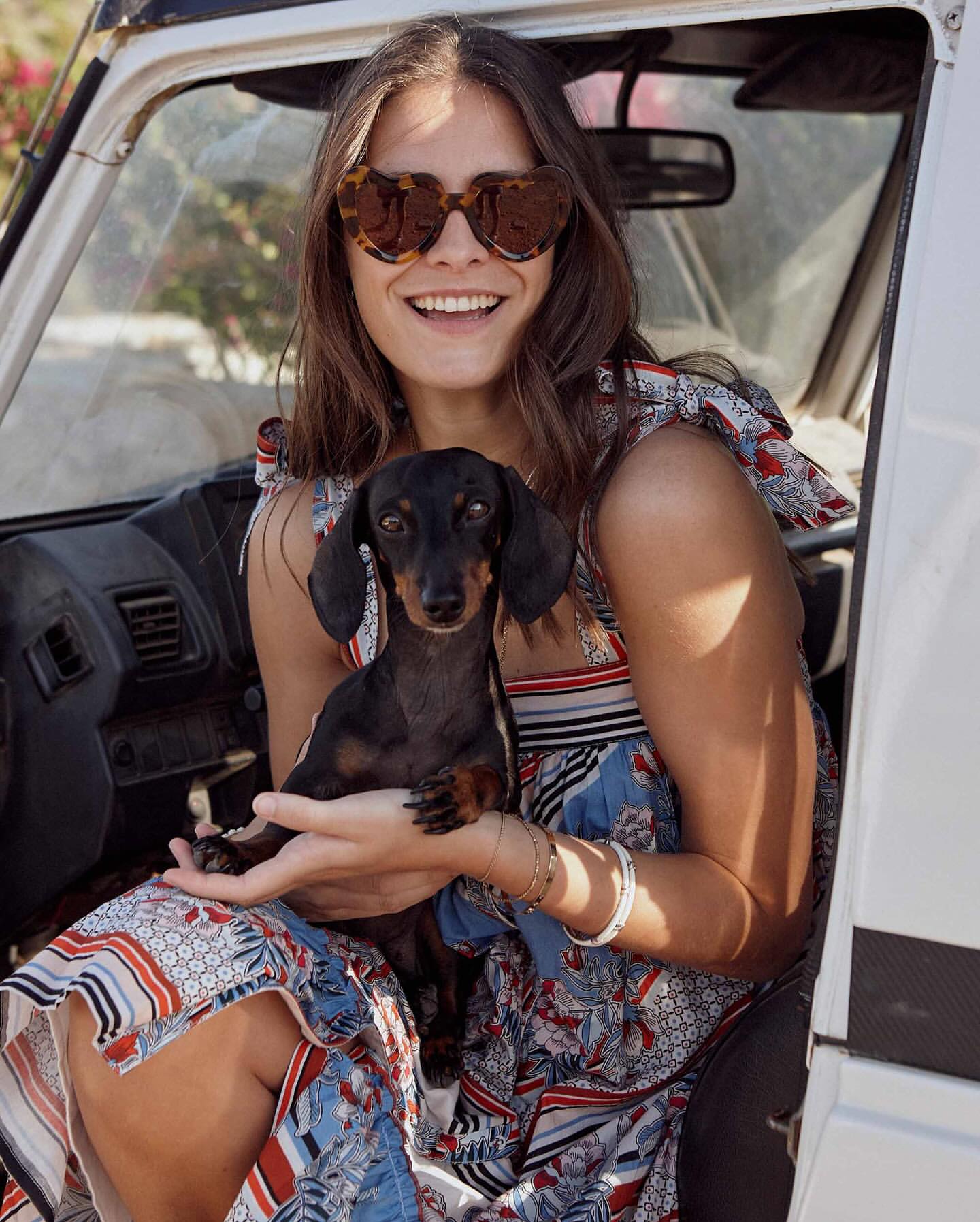 Smiling woman with dachshund in a white car. Wearing sunglasses and a patterned dress. Outdoor setting, daytime.
