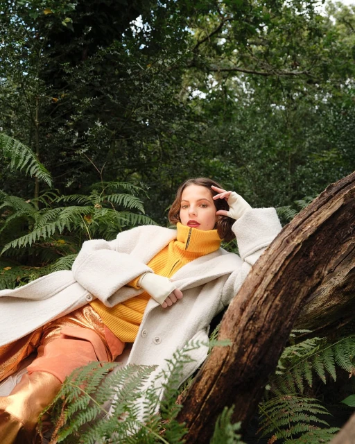 Stylish woman in a cream coat and orange pants poses reclining against a tree in a forest setting, captured in natural light.