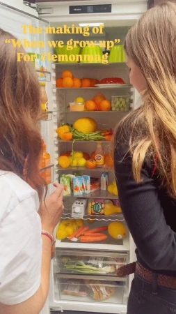 Two women stand near a refrigerator filled with various fruits and vegetables, creating a colorful and vibrant composition.