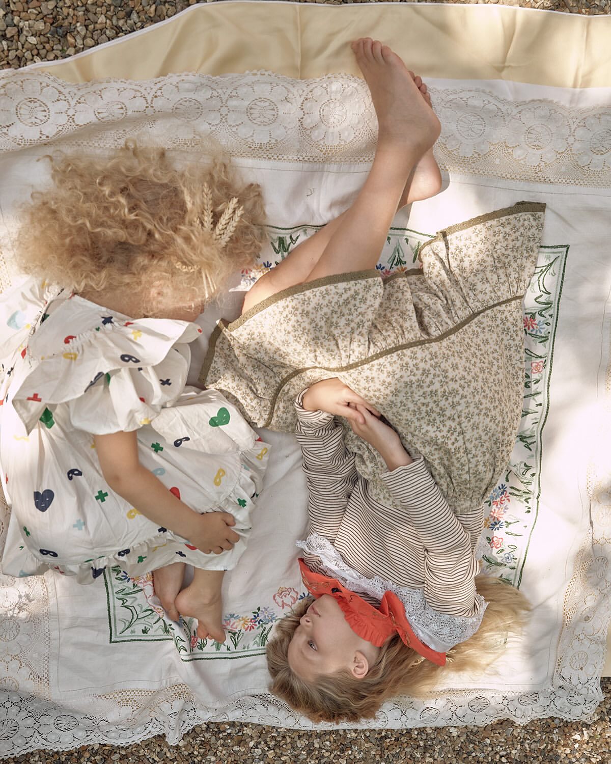 Two children lie on a patterned blanket outdoors in dappled sunlight. One wears a floral dress; the other wears a printed dress.