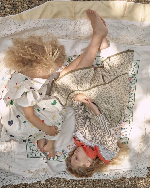 Two children lie on a patterned blanket outdoors in dappled sunlight. One wears a floral dress; the other wears a printed dress.