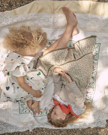 Two children lie on a patterned blanket outdoors in dappled sunlight. One wears a floral dress; the other wears a printed dress.