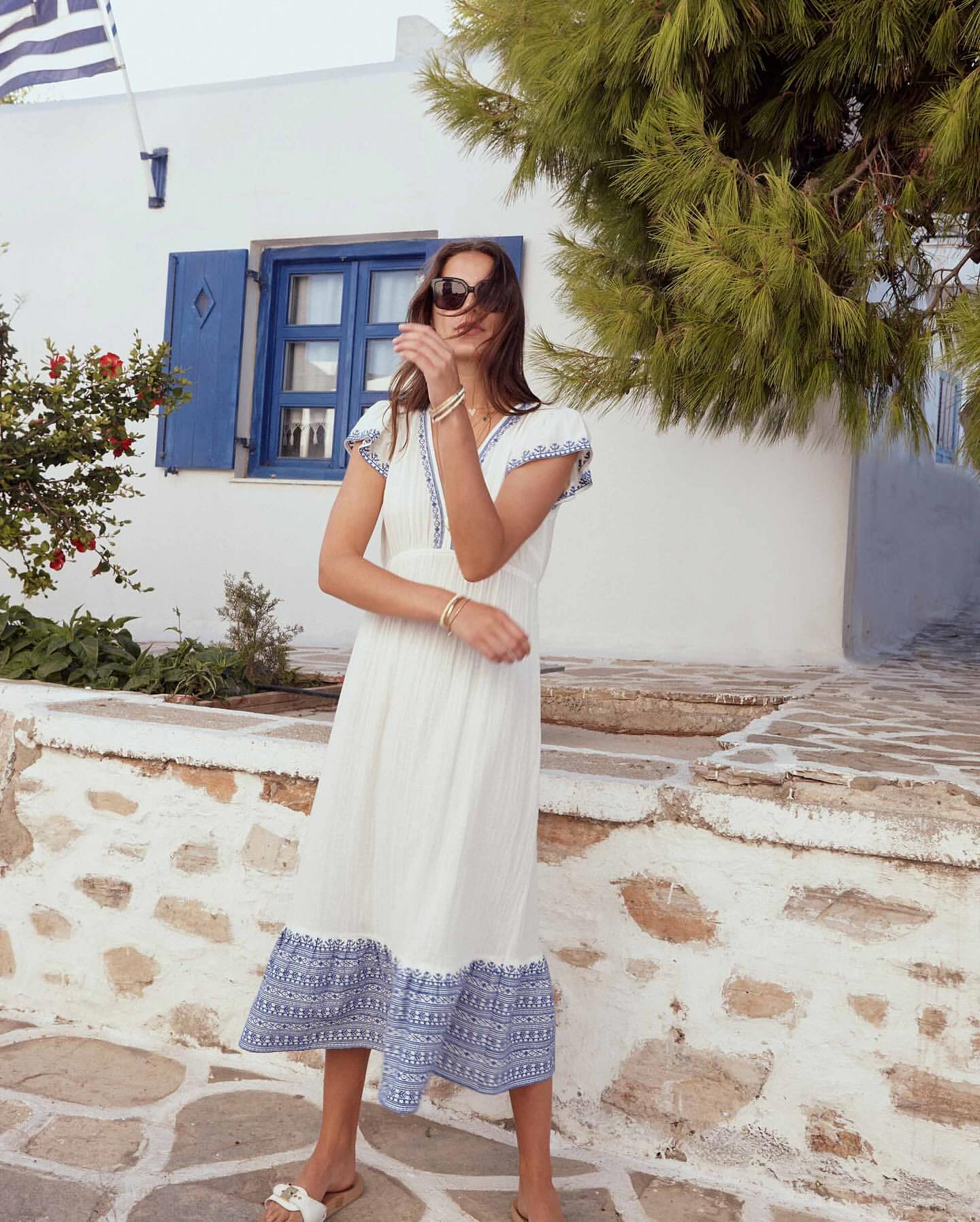 Adult woman in a white sundress with blue embroidery stands in front of a white building with blue shutters, Greek flag on top.