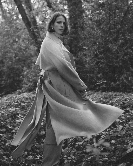 Monochrome portrait of a woman in a long coat with tie belt, standing in a forest clearing with scattered leaves.