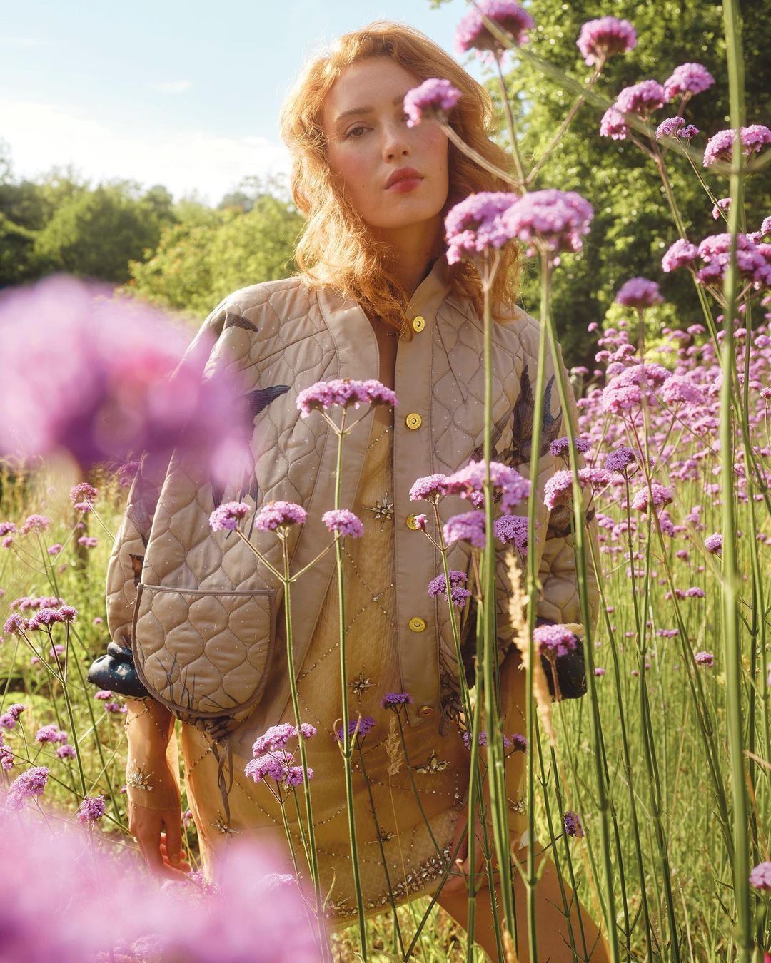 Woman in a quilted jacket and dress stands amongst purple flowers in a sunny field, looking at the camera with a neutral expression.