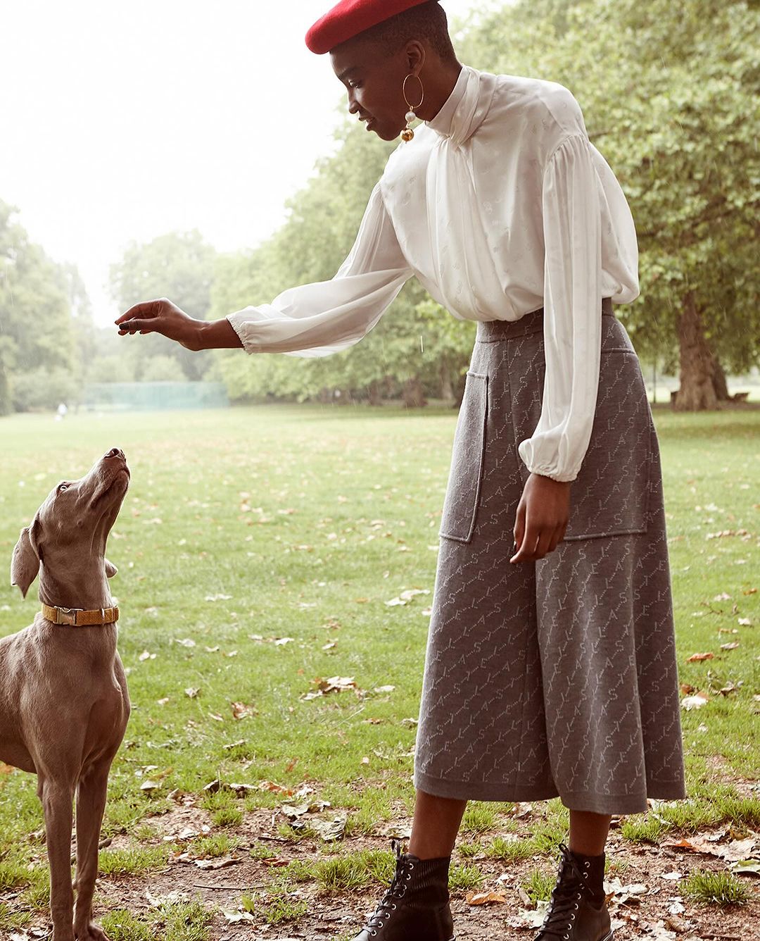 Elegant woman in beret with Weimaraner dog outdoors. Stylish female in white blouse and grey skirt interacts with her pet on green grass.