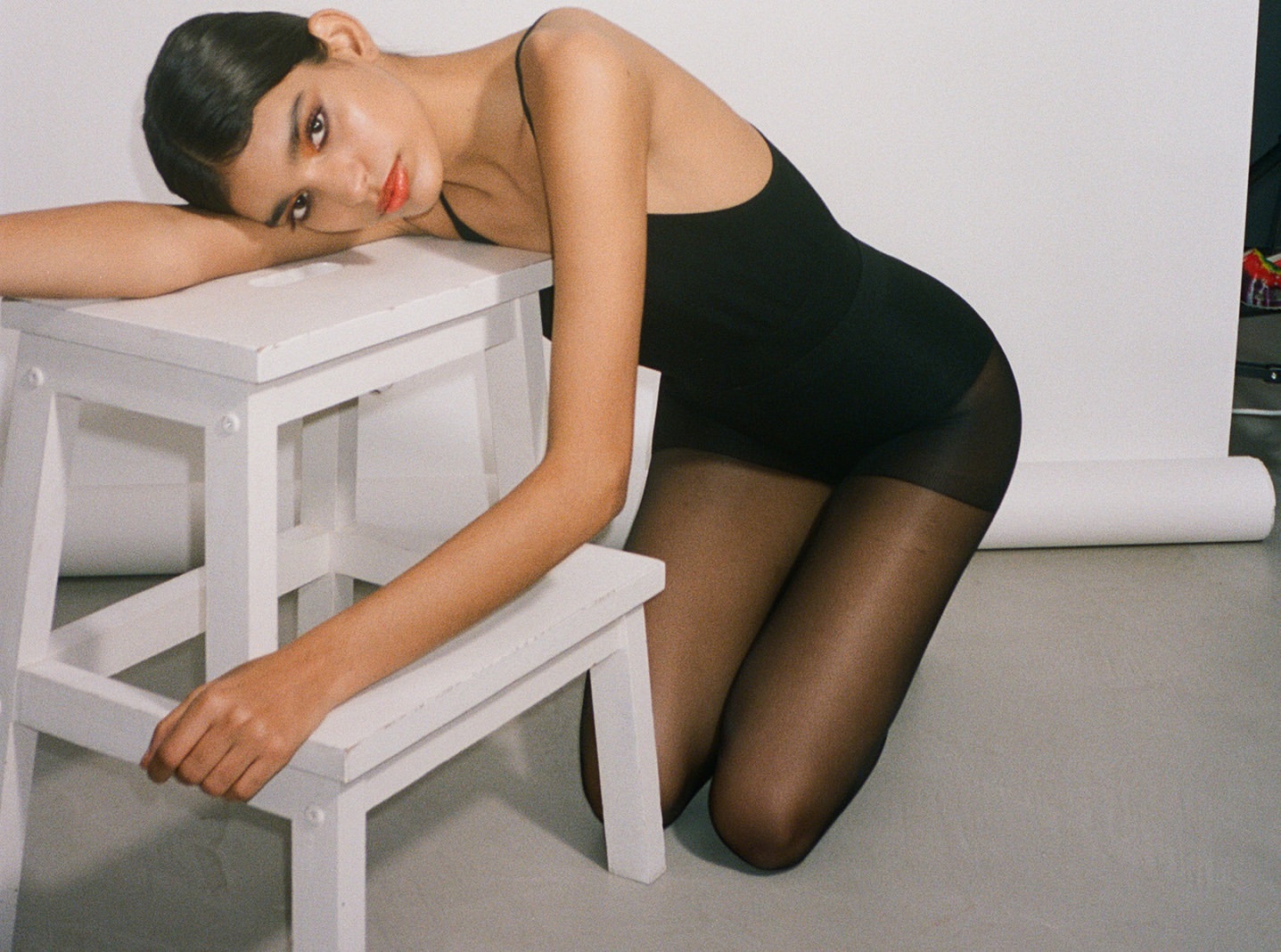 Woman in black bodysuit and tights poses leaning on a white step stool in a photo studio with a white backdrop and neutral lighting.