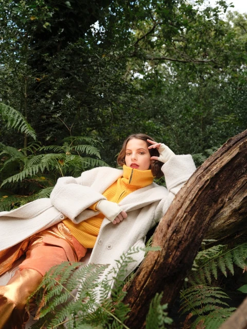 Woman in coat and yellow sweater reclining on a log in a forest setting, surrounded by ferns and greenery, looking at the camera.