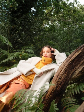 Woman in coat and yellow sweater reclining on a log in a forest setting, surrounded by ferns and greenery, looking at the camera.