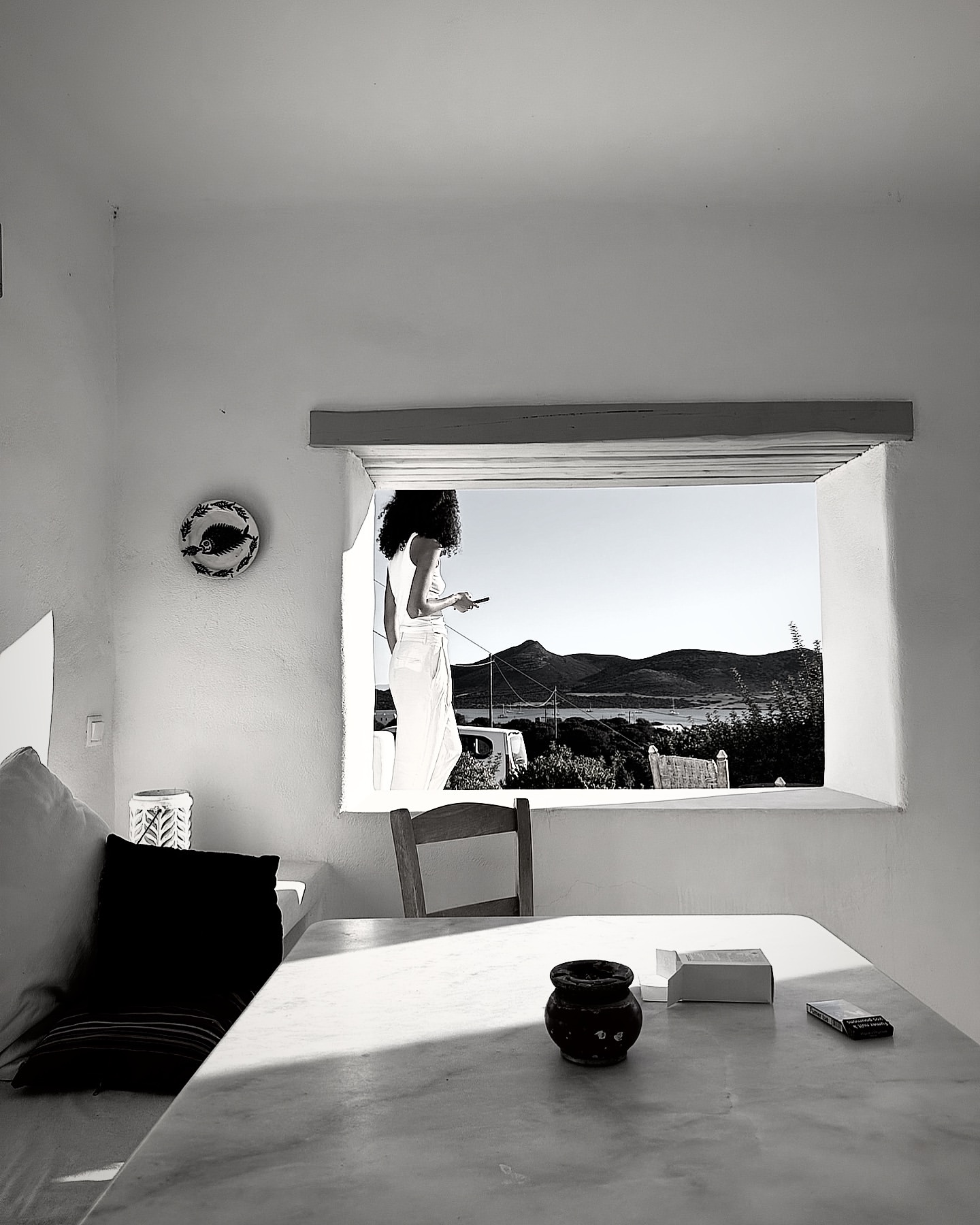 Monochrome view of a woman in a white robe using a smartphone by a window overlooking mountains and a sea inlet.
