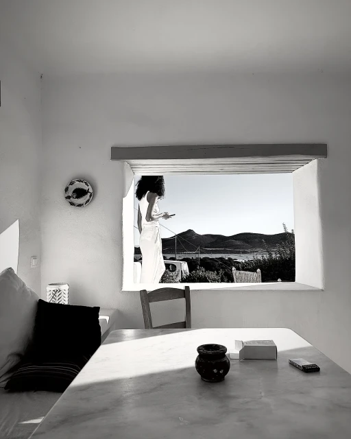 Monochrome view of a woman in a white robe using a smartphone by a window overlooking mountains and a sea inlet.