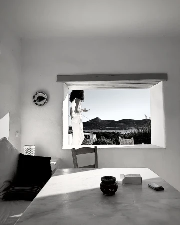 Monochrome view of a woman in a white robe using a smartphone by a window overlooking mountains and a sea inlet.