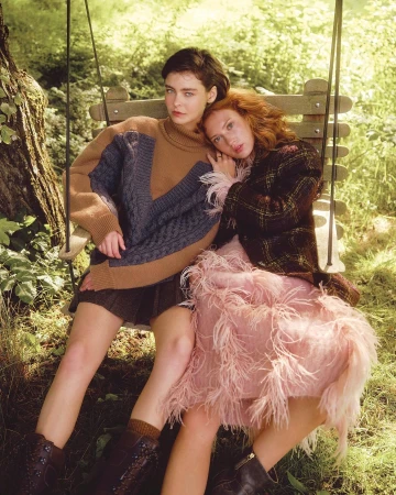 Two women seated together on a wooden swing suspended outdoors amidst lush green trees and foliage.