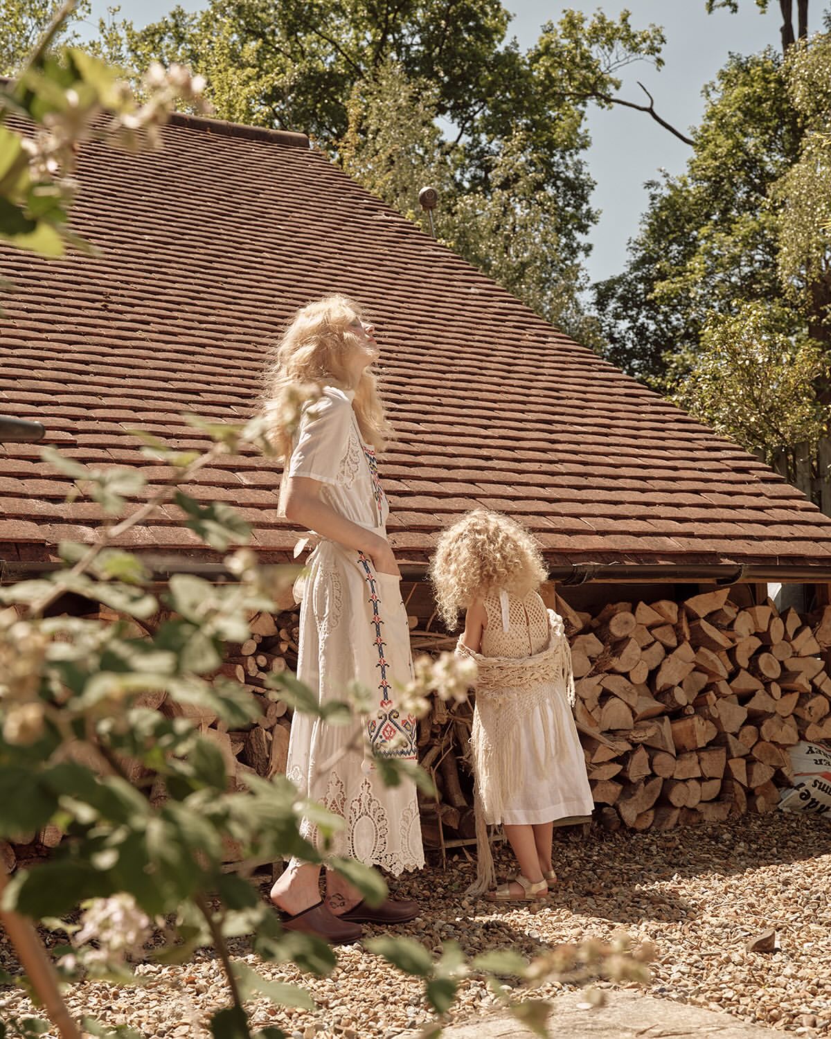 Woman and child stand near a wood pile next to a brown roofed structure. Both wear light colored dresses in an outdoor, sunlit scene.