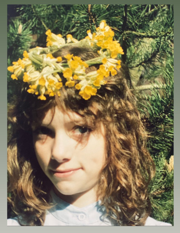 Portrait of a young girl with curly hair and a floral wreath, standing outdoors with a forest background, capturing a natural and youthful aesthetic.