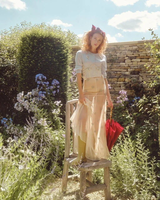 A young adult woman stands on a wooden stool in a sunlit garden, wearing a light-colored top, a sheer skirt, and holding a red umbrella.