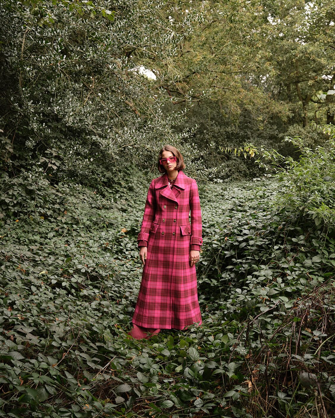 Woman in plaid coat standing in forest. Eye-level medium shot highlights fashion and natural environment with dense green foliage.