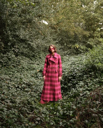 Woman in plaid coat standing in forest. Eye-level medium shot highlights fashion and natural environment with dense green foliage.