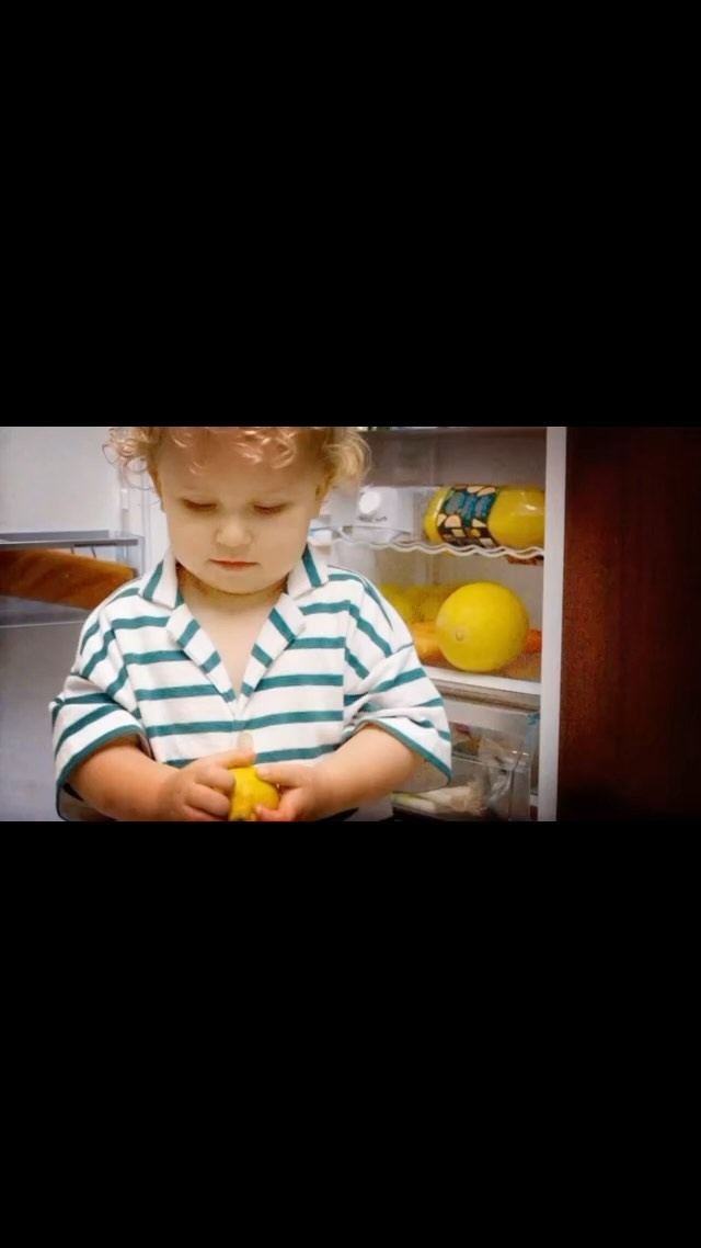 Child standing in front of open refrigerator, holding a lemon, wearing a striped shirt in an indoor setting with bright lighting.