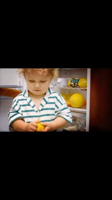 Child standing in front of open refrigerator, holding a lemon, wearing a striped shirt in an indoor setting with bright lighting.