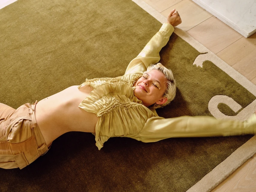 Woman with platinum blonde hair and gold makeup lying on green decorative rug with arms outstretched, wearing tan pants and silk top.