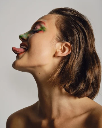Profile portrait of a woman with wet-look hair and colorful face paint sticking her tongue out against a light background.