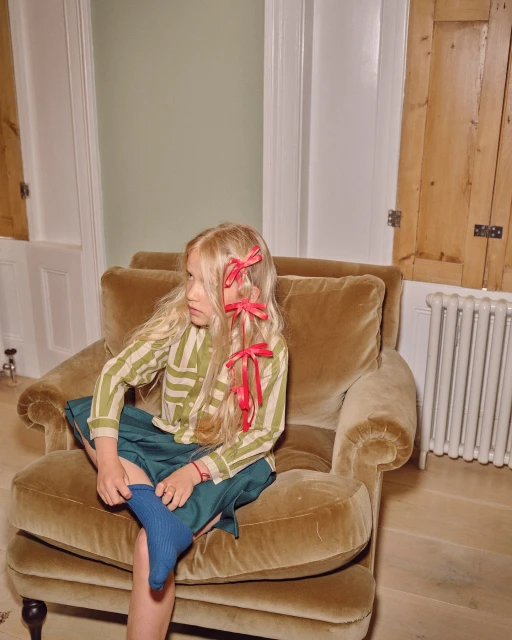 A child with blonde hair wearing a striped shirt sitting on a gold velvet chair with a skirt and one sock on in an indoor setting.