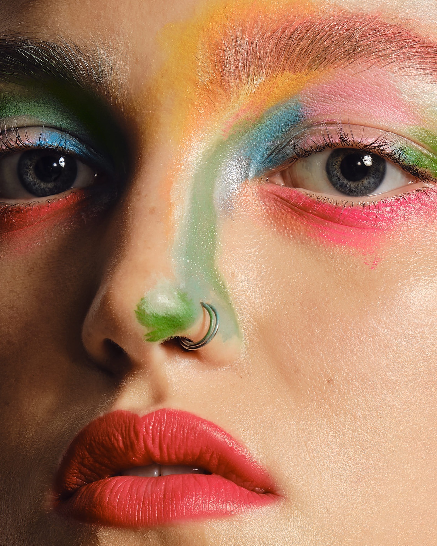 Close-up of a woman's face with colorful makeup in shades of blue, green, pink, and yellow; facial piercings; and red lipstick.