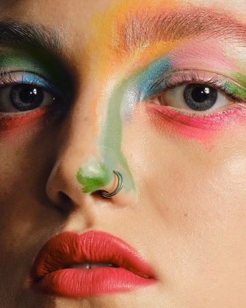 Close-up of a woman's face with colorful makeup in shades of blue, green, pink, and yellow; facial piercings; and red lipstick.