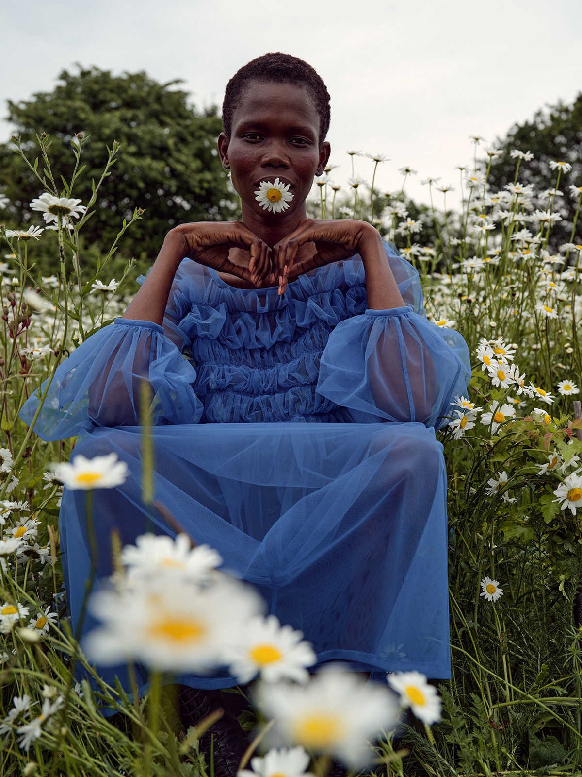 A dark-skinned woman with a daisy in her mouth sits in a field of daisies, wearing a blue dress with balloon sleeves.