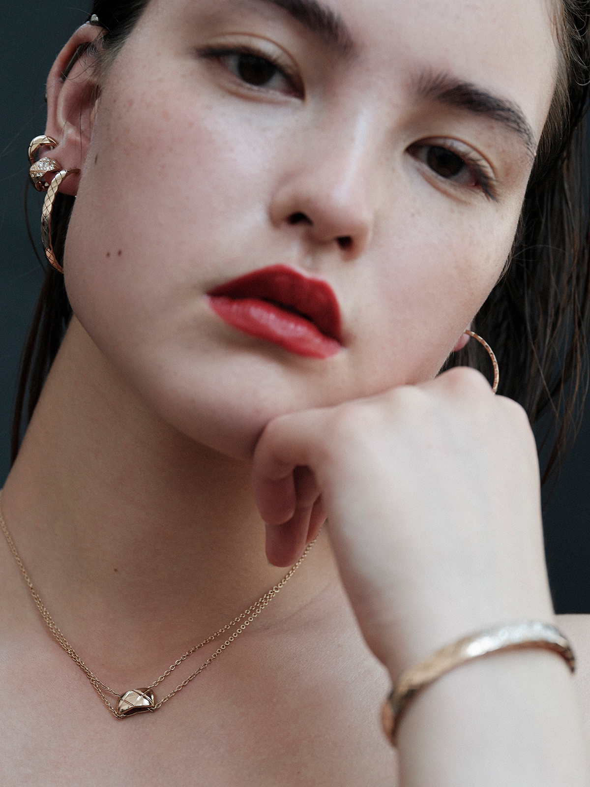 Close-up studio shot of a female model with red lipstick wearing gold hoop earrings, necklace, and bracelet against a dark background.