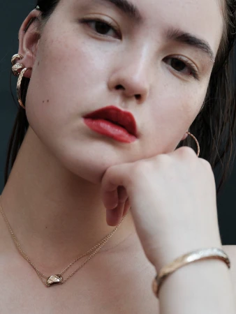 Close-up studio shot of a female model with red lipstick wearing gold hoop earrings, necklace, and bracelet against a dark background.