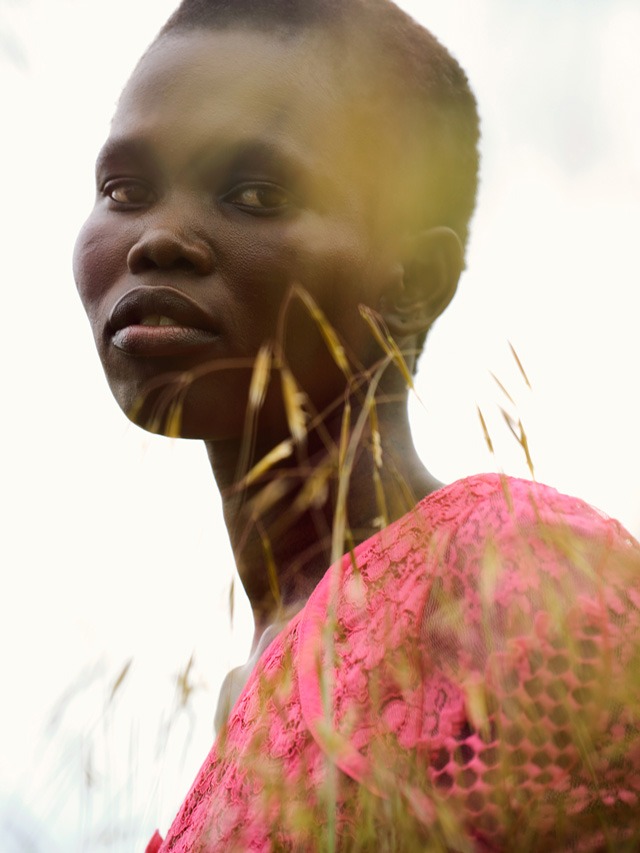 Close-up of a black woman with short hair wearing a pink lace dress, gazing at the camera, with blurred grass in the foreground.