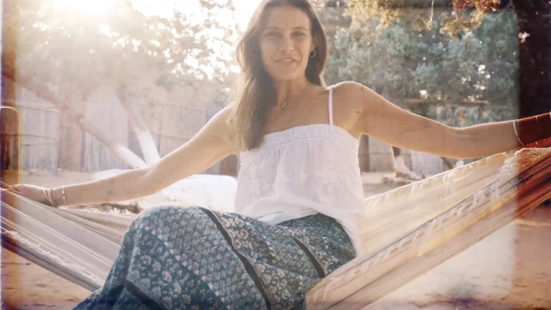 Woman in white camisole and patterned skirt resting in a hammock outdoors during sunset