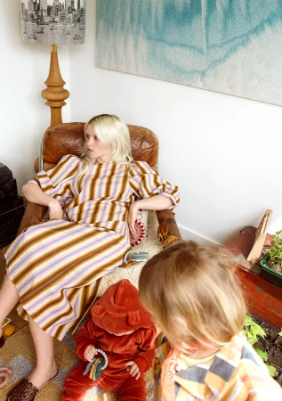 Woman in striped dress sits in a leather chair while a toddler in a bear suit plays on a rug in a well-lit, home interior setting.