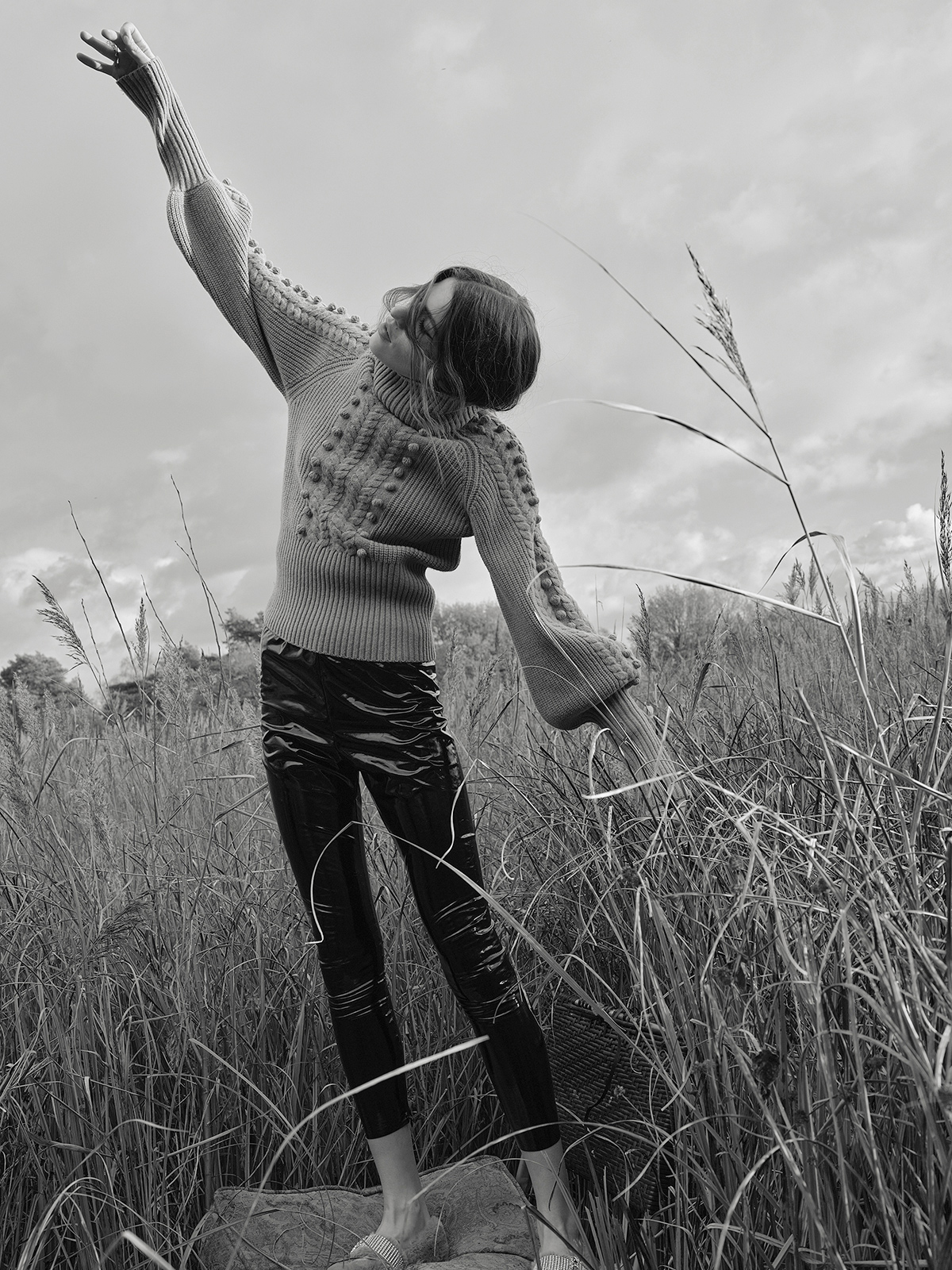 Monochrome fashion shot of a woman in sweater and leggings in a grassy field, arms raised, shot from a low angle against a cloudy sky.