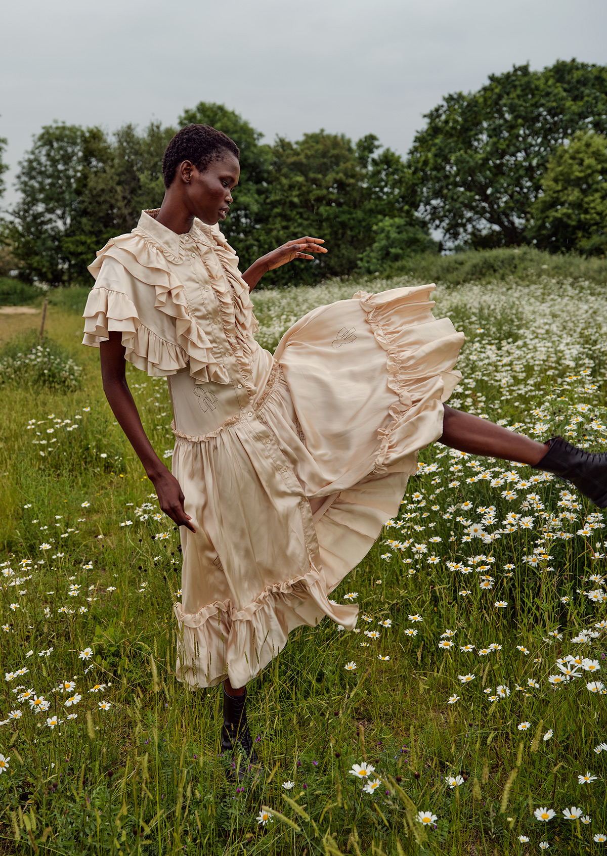 An adult female model wearing a ruffled dress in a grassy field with white flowers, trees and cloudy sky in the background.
