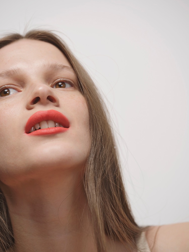 Close-up portrait of a young woman with tan hair and bright red lipstick looking up against a plain white background, fashion and beauty shot.