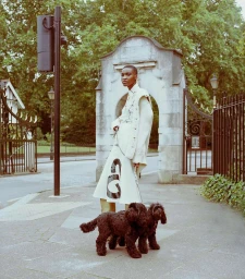 Model standing on a sidewalk holding leashes of two black dogs in front of an ornate stone archway and black iron gates.