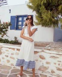 Woman in white dress with blue embroidery wearing sunglasses standing outdoors by a white building with blue shutters