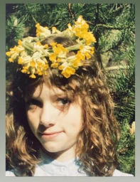 Young person with curly brown hair wearing a bright yellow flower crown outdoors with pine tree branches in the background