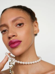 Close-up beauty portrait of a woman wearing a pearl necklace with a number five charm and magenta lipstick against a white background