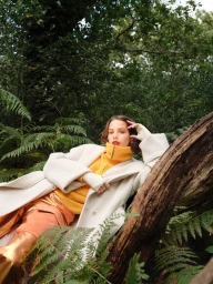 Woman in a yellow turtleneck and white coat reclining against a fallen log in a lush forest