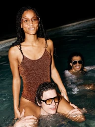 Three young adults swimming in a pool at night, with a woman sitting on a man's shoulders wearing a glittery brown swimsuit.