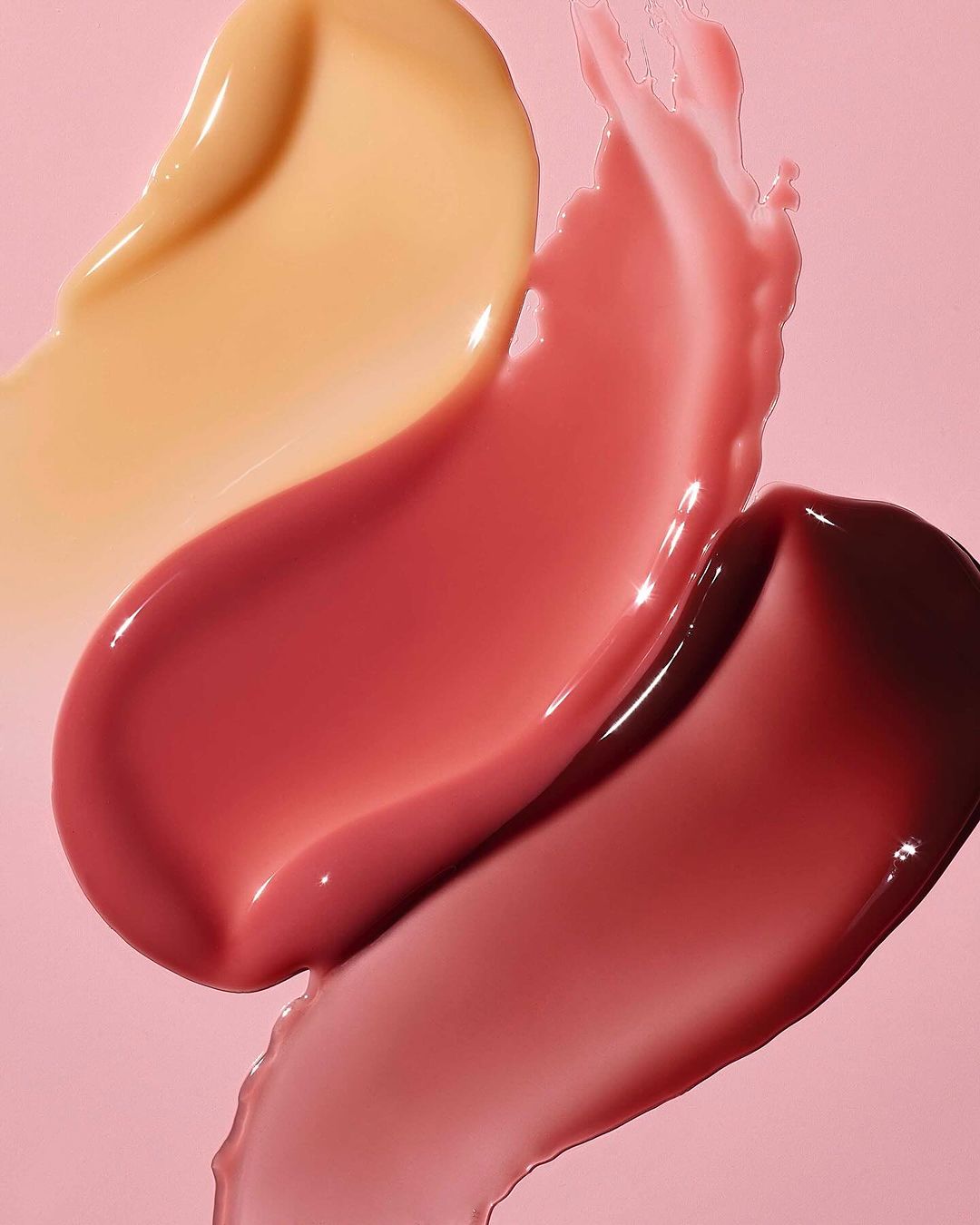 Macro photo of lipstick and lip gloss smears in peach, red, and berry tones against a light pink backdrop, beauty product textures.