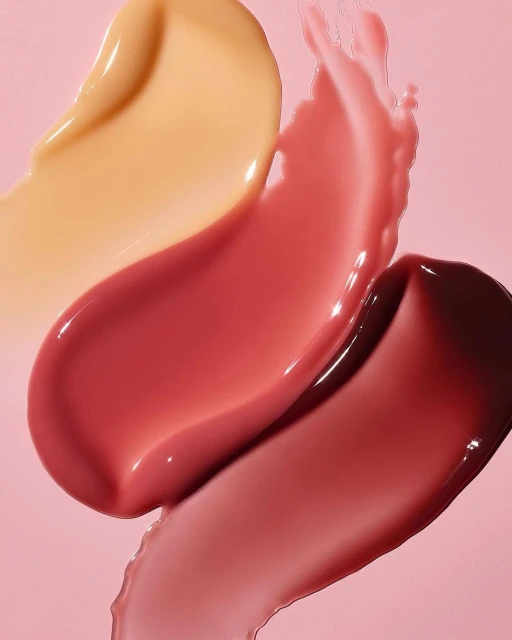 Macro photo of lipstick and lip gloss smears in peach, red, and berry tones against a light pink backdrop, beauty product textures.
